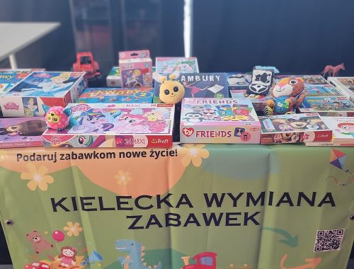 Stół z grami i zabawkami przygotowanymi do Kieleckiej Wymiany Zabawek, ozdobiony kolorowym banerem akcji.